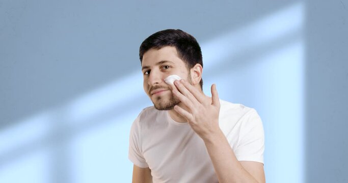 Slow motion photo of bearded man applying moisturizer or face serum with two fingers in bathroom on light blue background. Man smiling nicely at camera. The concept of professional care products