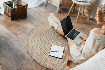 blonde woman in sweater and knitted socks using laptop in modern apartment.