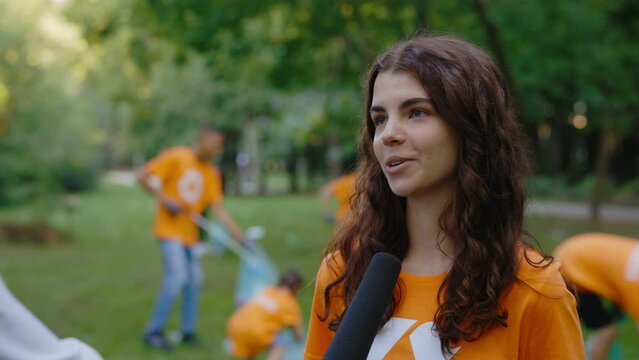 Young Caucasian Female Eco Activist is Giving the Interview to Journalist Standing in the Public Park. A Group of Volunteers Collects Trash in the Background. Save the Planet. News