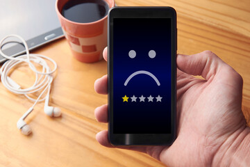Male hand holding mobile phone with sad face and one star rating. Very unhappy customer giving...