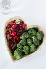 Colorful heart shape from various fruits and vegetables with glass of fresh water isolated on white background. Vertical shot, top view. Love for healthy plant-based food concept. 