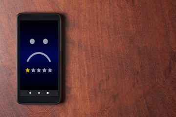 Mobile phone with sad face and one star rating on wooden table with copy space. Very unhappy...