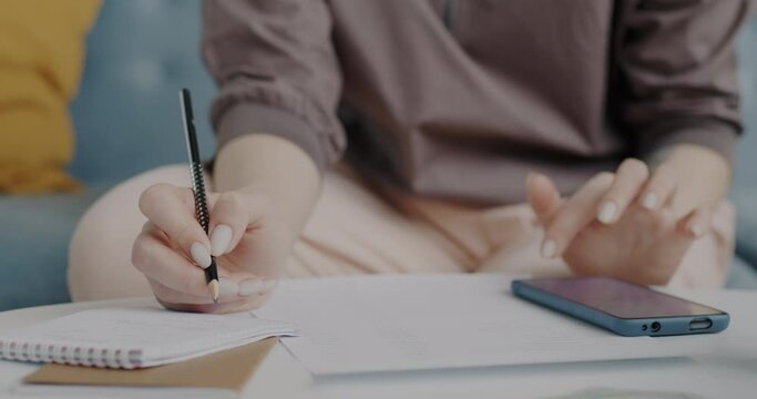 Close-up of female hands using smartphone and writing notes on paper indoors at home. Modern technology and business paperwork concept.