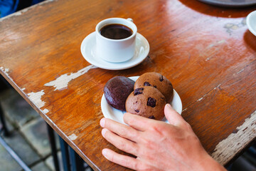 A man's hand holds a chocolate chip cookie against the background of a mug of Americano coffee. - obrazy, fototapety, plakaty