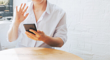 A woman in a white shirt waves her hand and holds the phone at the table. Stylish banner of a freelancer working from a cafe. Online video communication and training by phone