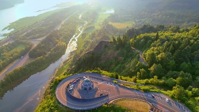 Aerial view at golden hour during sunrise-a slow flying forward over and past Vista House showing Mirror Lake, the Columbia River and a train traveling East. The sun’s rays and mist create a soft glow