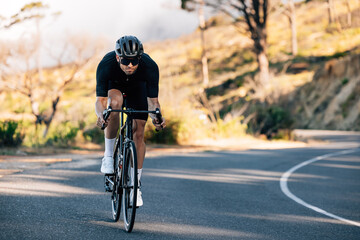 Professional cyclist in black sports attire going down a hill. Cyclist in a helmet and glasses riding on a bicycle outdoors.