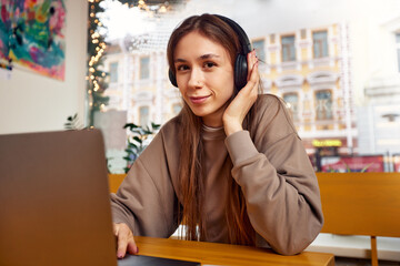 A young girl working on a laptop with a phone and a cup of coffee poses for the camera in a coffee shop against the background of talking people.