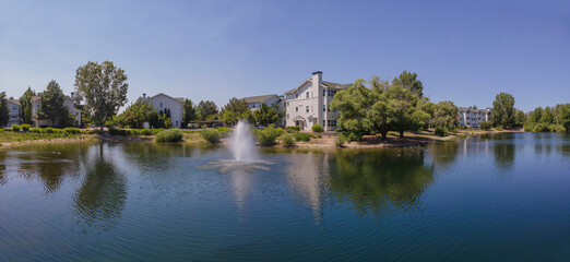 Fototapeta na wymiar Beautiful apartments with view of a small lake with fountain in Boise Idaho. Scenic landscape of rental properties overlooking a reflective body of water against blue sky.