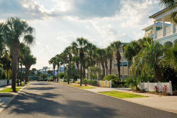 Street in a residential area with fenced houses and sidewalks along the trees in Destin, Florida. There is an asphalt road in the middle against the sky.