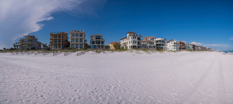 Panorama of Destin beach houses in Florida with fenced sand dunes at front. White sand and dunes at the front of the large houses with giant clouds in the blue sky background.