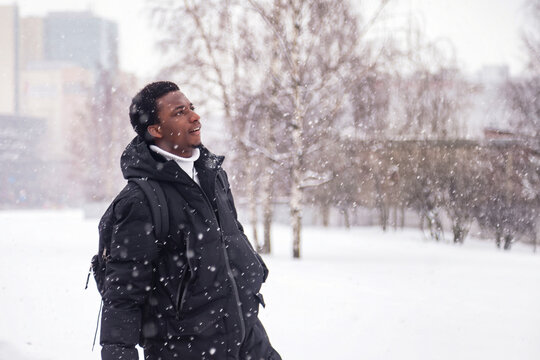 African american man standing in snowy russian winter at urban background, looking up away. Portrait black man walking of city, falling snow outside. Lifestyle wintertime concept. Copy ad text space
