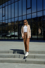 Young woman with visual disability walking near business center with cane stick in hands. Caucasian lady in smart casual wear checking safety direction outdoors.