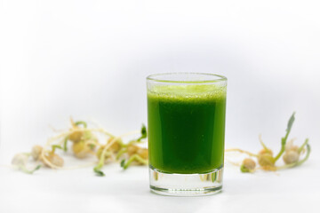 Green Wheatgrass juice in a transparent glass. On a white background with blurry sprouts
