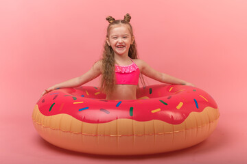 A little child girl in a swimwear suit lying on a donut inflatable circle. Pink background. Summer...