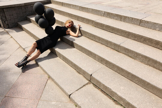 Beautiful woman in black dress with dark balloons lies on stairs in park. Art shooting from unusual angles. High quality photo
