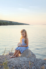 A beautiful girl with long blond hair sits on the shore and admires a large sea shell.