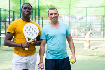 Portrait of positve men, african-american and caucasian, padel tennis players standing on outdoor court.