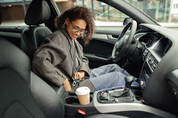 Smiling woman driver in eyeglasses fastens her seat belt before starting car. Driving safety concept - obrazy, fototapety, plakaty