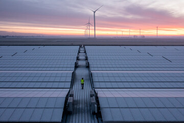 A construction working walks through a solar field with the solar panels covered in snow. They don’t produce any power like this. Wind turbines for power production are seen at the horizon.