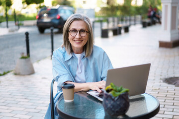 Happy and content Caucasian mature woman with long grey hair, seated at a street cafe with a notebook. She appears relaxed and enjoying her leisure time while staying connected with the world through