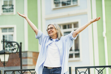Mature woman standing in old city, smiling and raising her hands to sky, expressing happiness and...