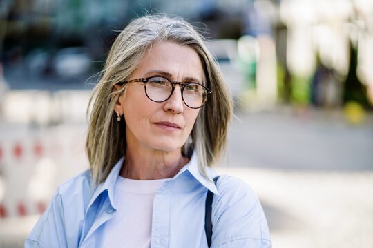 Sad mature woman with silver hair and glasses in a European city, portraying feelings of loneliness, depression, and despair in the context of aging and mental health issues in seniors.