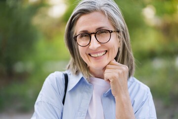 Happy mature woman with silver hair and glasses smiling outdoors, showcasing the beauty of aging...