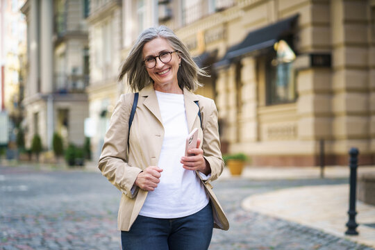 Joyful mature woman holds her phone while standing in old European city. This image captures the concept of happiness in the elderly.