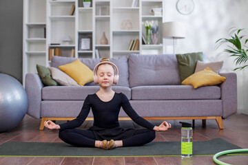 Child doing yoga exercise in room. Healthy lifestyle. Girl listens to music in headphones.