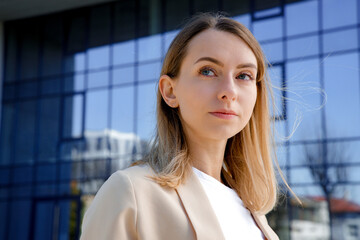 Portrait of confident young woman with blond hair looking at camera outdoor. Serious female standing near glass wall of office building. Concept of people and emotions.