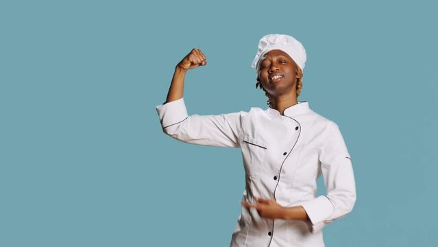 Powerful woman cook flexing arm muscles on camera, showing power and body strength in studio. Confident happy chef in cooking apron being good at gastronomy and cuisine industry.