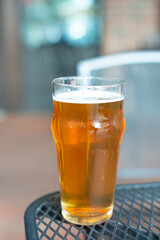 View of a glass of sour flavored beer with its head covered in froth. The liquid has an orange tint. There's a plastic cup cover on the beer on a composite plastic table top at a microbrewery. 
