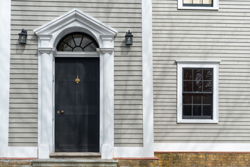 The exterior of a vintage building with beige colored narrow clapboard cape cod siding. There's a black wooden door with a thick white decorative trim. A half circle transom window hangs over the door - obrazy, fototapety, plakaty