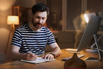 Home workplace. Man taking notes while working with computer at wooden desk in room at night