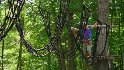 A little boy is enjoying an exciting adventure on a rope obstacle course in a lush forest. He is wearing a helmet and a safety harness as he navigates through the challenging course, showing bravery