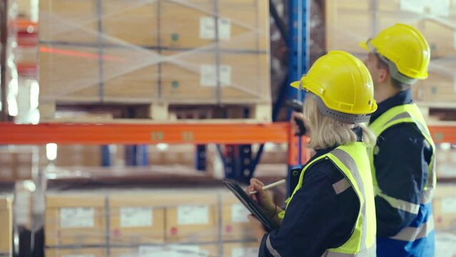 Warehouse workers holding digital tablet checking inventory management packaging boxes. Two staff wearing vest and safety helmet and walking count the box goods on shelves at storehouse