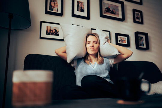 Woman Sitting on a Sofa Covering her Ears with Pillows. Unhappy lady suffering from a headache due to loud neighbors and thin walls
