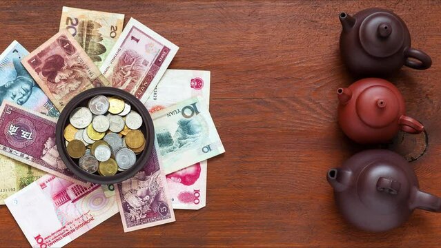Flat lay of Chinese Yuan coins in a cup on paper Yuan banknotes and next to clay chinese teapots with green tea on wooden background. Asian trade concept, green tea value. Top view, close-up, mockup