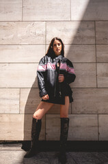 Urban chic: young brunette model wears leather jacket and long boots in big stone building with morning sunlight