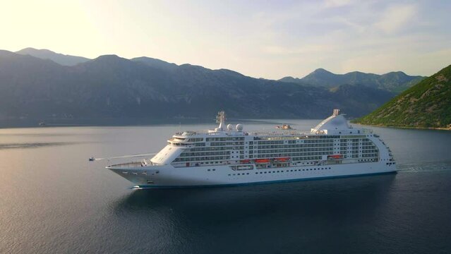 The drone captures a stunning aerial view of a white cruise ship leaving the Boka-Kotor bay in Montenegro during sunset. The calm sea and surrounding mountains create a serene atmosphere, making it a