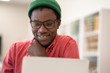 Cheerful happy African guy student looking at laptop screen while studying in college library, selective focus. Positive young black man enjoying e-learning, watching interesting webinar on computer