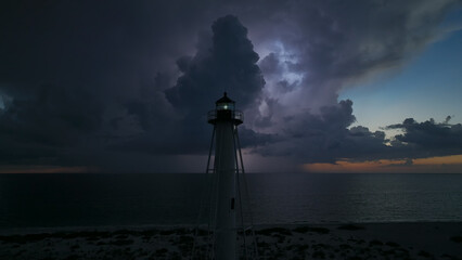 White tall lighthouse on sea shore with blinking light at stormy night for commercial vessels navigation. Thunderstorm with lightnings over ocean water posing danger for ships