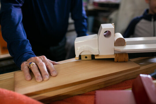 old caucasian man's wrinkled hands on pieces of wood as he makes wooden trucks in woodworking shop