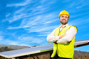 Portrait of Young indian man technician wearing yellow hard hat standing near solar panels against blue sky.Industrial worker solar system installation, renewable green energy generation concept.