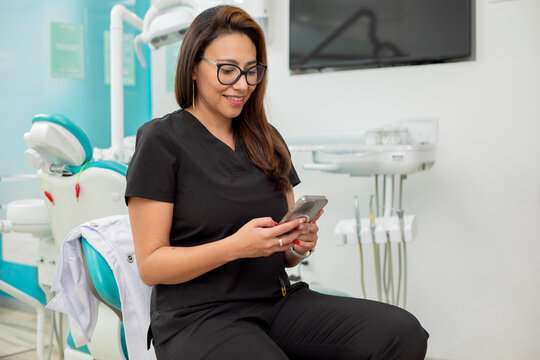 Young Latina dentist sitting in her office, checking her cell phone - Powered by Adobe