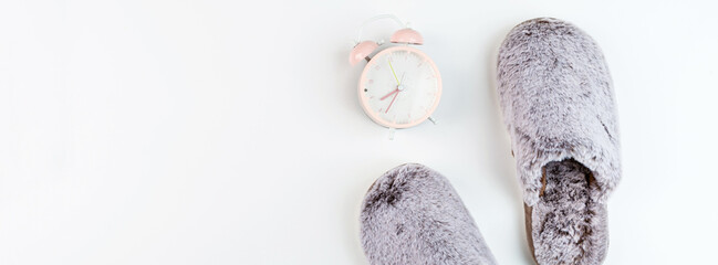 Flat lay with Soft fluffy slippers gray color and pink alarm clock isolated on white background....