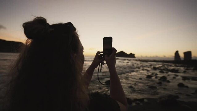 Woman takes picture with phone by rocky Azores coast at dusk, close-up