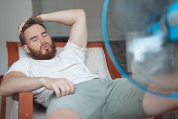 Obraz premium Young bearded man using electric fan at home, sitting on couch cooling off during hot weather, suffering from heat, high temperature