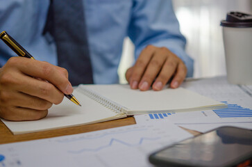 Businessman using pen writing note book planning journal and analysis information statistics document on desk.